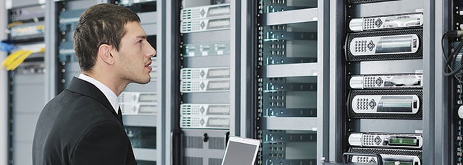 Male information technology professional standing in a server room with an open laptop while examining network hardware