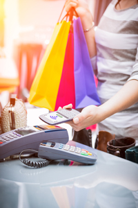 Close-up of a woman's hand using a smartphone to make a contactless payment at a store checkout counter with colorful shopping bags and POS terminal in the background.