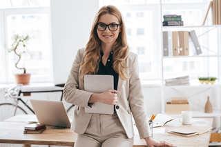 Professional woman in glasses holding a tablet, standing in an office with a laptop and paperwork on the desk.