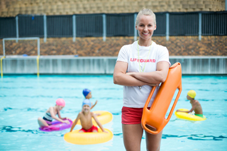 Female lifeguard with rescue tube standing by outdoor swimming pool with children playing in water in the background