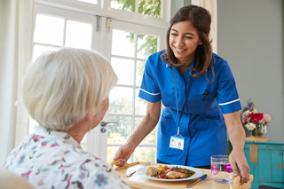 Healthcare worker in blue scrubs serving meal to elderly woman at care home, smiling as they interact in a bright, airy dining room.
