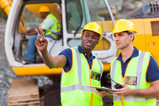 Two construction workers in safety vests and helmets discussing at a construction site with heavy machinery in the background.