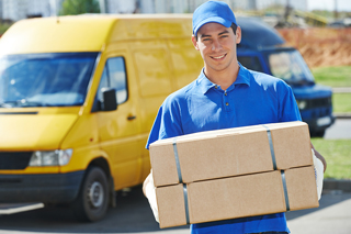 Delivery man in blue uniform smiling and carrying cardboard boxes with yellow van in background.