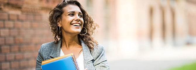 Young woman in a gray blazer smiling and standing outside a brick building while holding notebooks with both arms