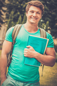 Young man smiling wearing a backpack and holding on to a notebook.