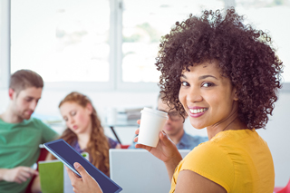 Young woman with curly hair holding a coffee cup and a tablet in a brightly lit office with colleagues working in the background.
