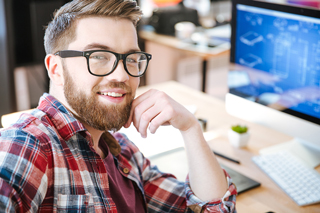 Smiling bearded man in glasses and plaid shirt sitting at desk with computer monitors displaying graphs