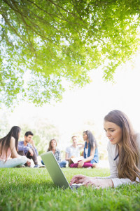 Young woman using laptop in park with friends in background sitting on grass under tree