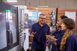 Senior male instructor discussing machinery with young female trainees in an industrial training facility.