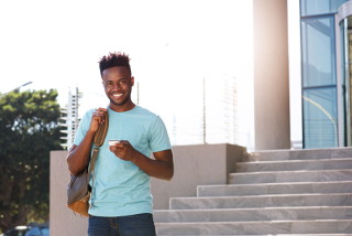 Young man with backpack smiling and using smartphone on sunny urban street