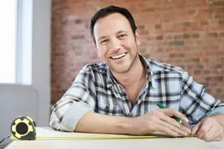 Man in plaid shirt sitting at desk with measuring tape, in a bright office with brick wall background.