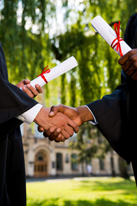 Two graduates in academic gowns shaking hands while holding diplomas, with a university campus setting in the background.