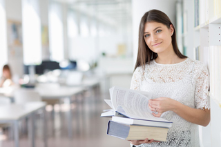 Smiling student holding textbooks beside a library bookshelf.