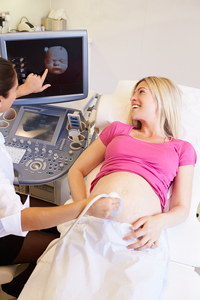 Pregnant woman smiling during ultrasound scan with technician pointing at baby's image on monitor in medical office.