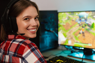 Young woman with headphones playing a video game, smiling at the camera in a room with gaming setup.