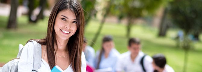 Smiling young woman standing outside with a gray backpack in front of a small group of sitting college students