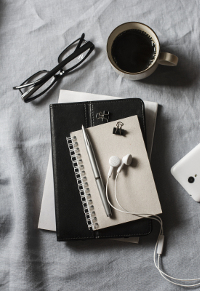 Stack of notebooks with eyeglasses, earbuds, and a cup of coffee on a textured gray cloth background