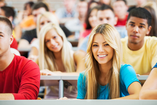 Diverse group of students in a classroom setting, focusing and smiling, with a young blonde woman in blue shirt at the forefront.