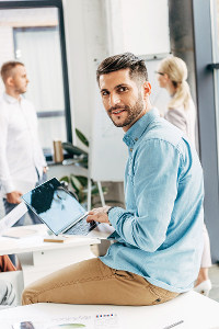 Man in blue shirt sitting at desk using laptop in office, looking over shoulder with a smile