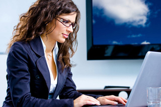Professional woman in business attire working on a laptop in an office setting with a monitor showing cloud imagery in the background.