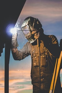 Welder in protective gear working with a welding torch against a sunset background