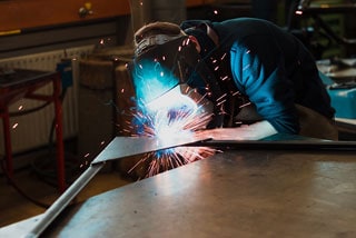 Welder wearing protective gear using welding torch on metal in workshop with sparks flying
