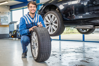 Male mechanic in blue coveralls holding a tire and a wrench in a car workshop with a black car lifted in the background