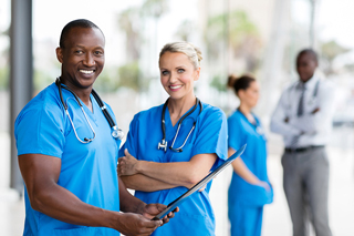 Two smiling healthcare professionals in blue scrubs with stethoscopes, holding a clipboard, standing in a hospital corridor with colleagues in the background.