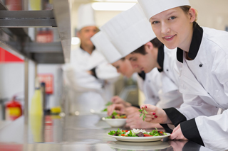 Chef in white uniform and chef's hat garnishing a dish in a professional kitchen with colleagues working in the background
