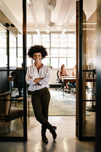 Confident young businesswoman standing in office doorway with colleagues working in background