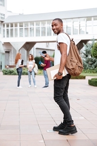 Smiling young man with a backpack holding books and looking back on a university campus with other students in the background.
