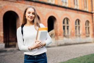 Smiling young woman holding books and wearing a backpack while standing in front of a brick building with arched doorways