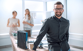 Man in business attire smiling at the camera with female colleagues discussing in the background in an office setting