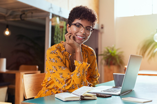Young woman with glasses sitting at a cafe with a laptop, books, and a calculator, wearing a yellow floral top.