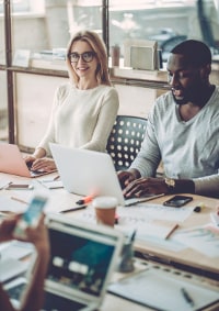 Two professionals, a woman and a man, working together on a laptop in a busy office environment.