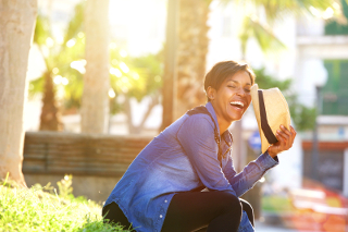 Happy young woman laughing and holding a book in a sunny park with palm trees.