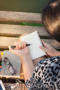 Woman sitting on wooden dock writing in blank notebook with glasses and earphones nearby
