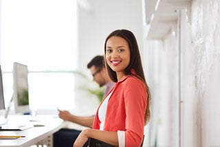 Young woman in red blouse smiling at camera in a bright office setting with colleagues working in the background
