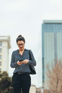 Woman in striped blouse and glasses using smartphone while walking in urban environment with blurred buildings in background