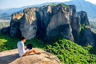 Man sitting on a cliff working on a laptop with a panoramic view of Meteora rock formations and green valley in the background.