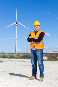 Engineer in safety vest and helmet standing with arms crossed in front of wind turbines on a clear day