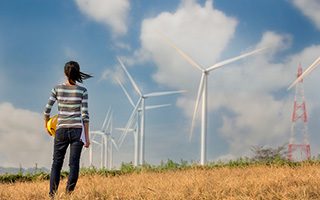 Woman with hard hat walking in a field towards wind turbines under a blue sky