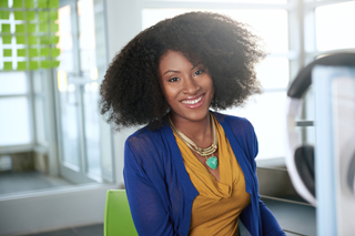 Woman with curly hair wearing a blue blazer and golden necklace sitting in an office.