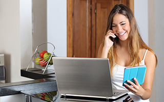 Woman using laptop and talking on phone in kitchen with books in hand