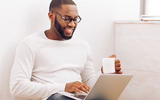 Male with glasses using laptop holding coffee mug in a bright room