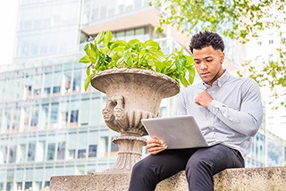 Young man using laptop while sitting on stone structure outdoors with building in background