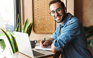 Man with glasses working on laptop and writing in notebook at a cafe table with plants nearby