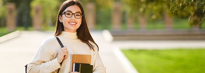 Female college student standing outdoors holding a set of books with one hand and the strap of a shoulder bag with the other